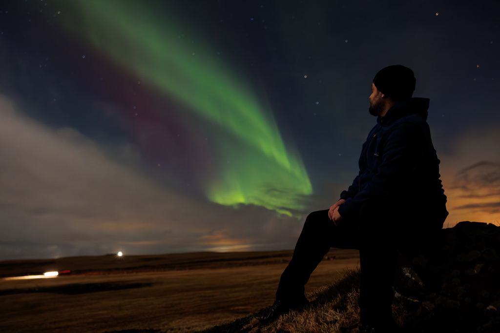 A lone traveller stands in the snow looking up at green aurora overhead