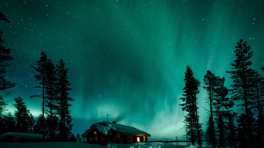 Green aurora above a lit wooden cabin and snow covered spruce forest in Finnish Lapland