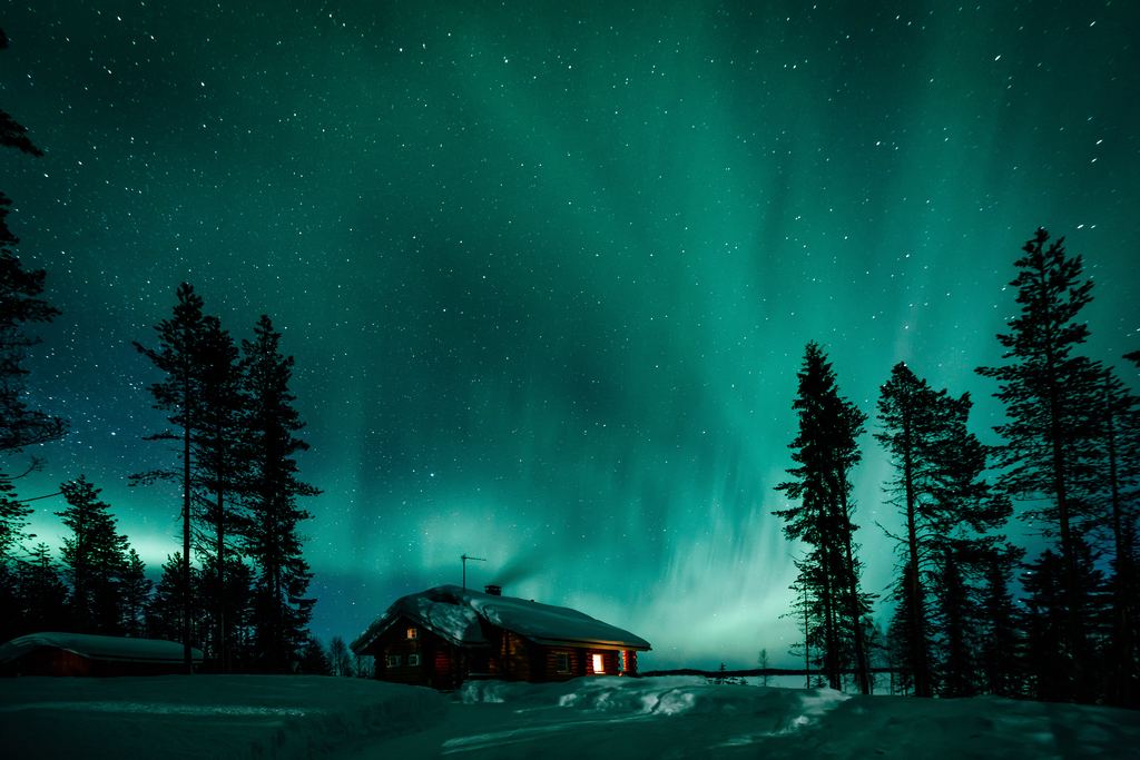 Green aurora above a lit wooden cabin and snow covered spruce forest in Finnish Lapland