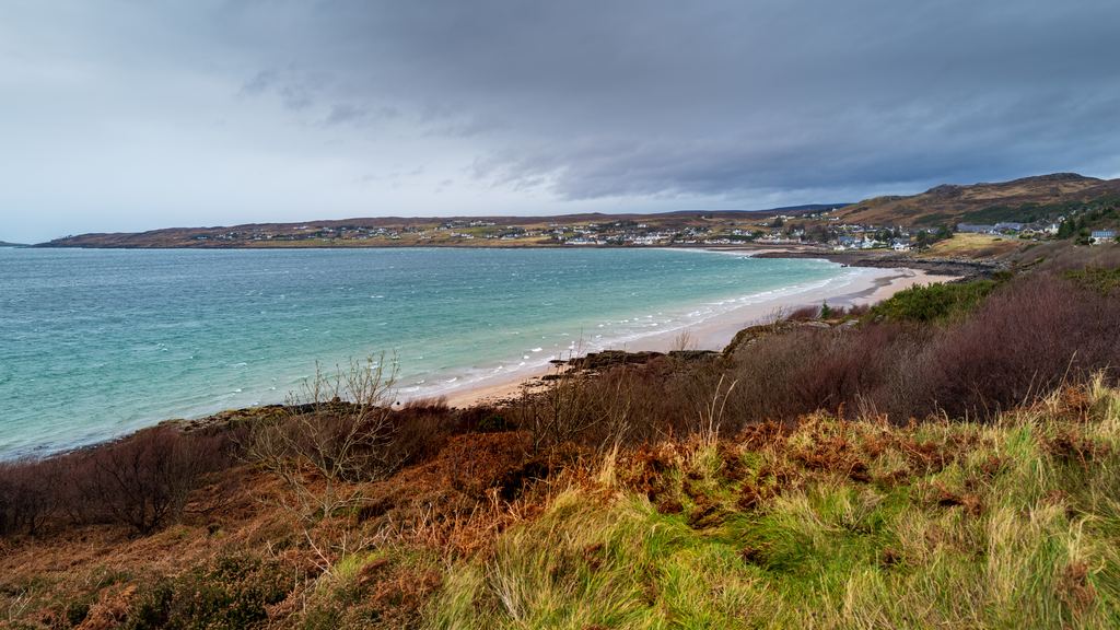 A bay with a pale sand beach, moorland and scattered white houses on the west coast of the Scottish Highlands under heavy grey cloud
