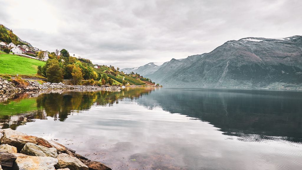 A calm Norwegian fjord under a low unbroken grey overcast, with mountains on both sides and a village on the near shore