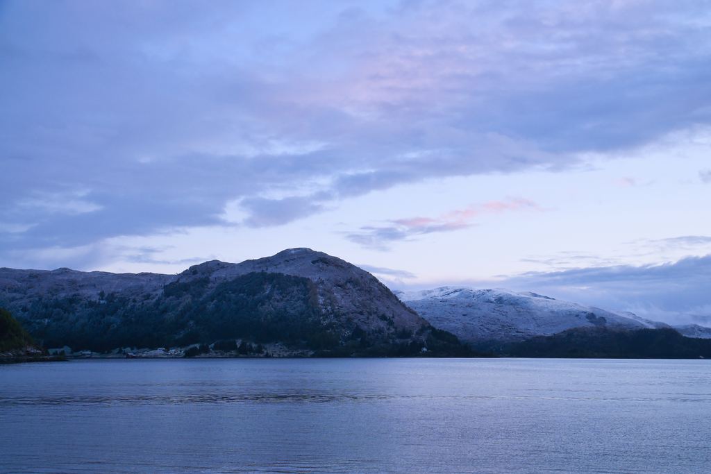Pale blue and pink twilight sky over a Nordic fjord and low mountains, with no darkness in the sky