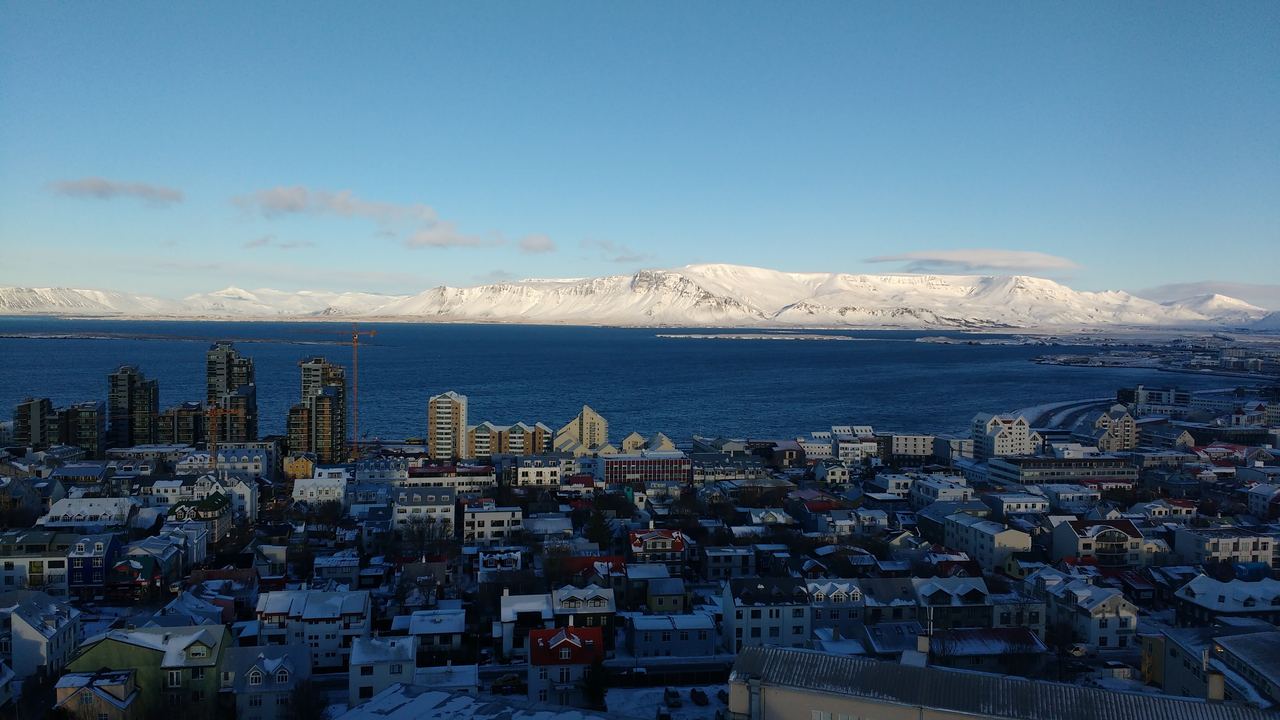 Aerial view of Reykjavík on the coast, with snow-covered mountains behind the city under a blue sky